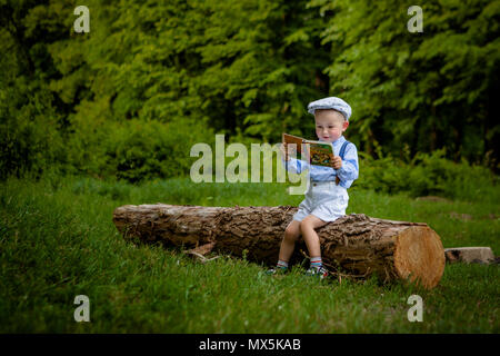 Ein litttle zwei Jahre alte Junge sitzt auf einem Baum und liest ein Buch. hildren Tag. Stockfoto