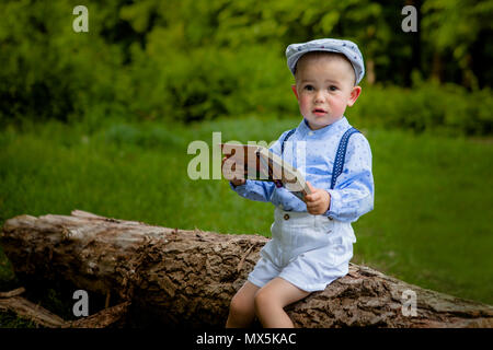 Ein litttle zwei Jahre alte Junge sitzt auf einem Baum und liest ein Buch. hildren Tag. Stockfoto