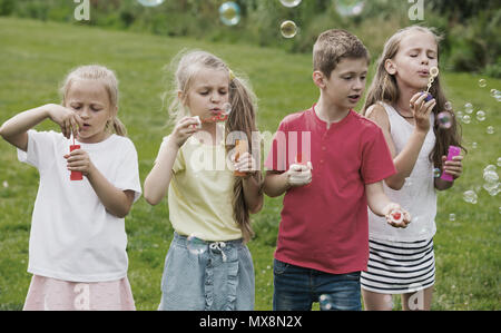 Verspielte Junge und Mädchen im Grundschulalter stehen auf grünem Gras und Seifenblasen Stockfoto