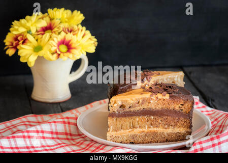 Appetitliche Scheiben von Kuchen mit Karamell und Schokolade füllen, mit gelben Daisy - wie Blumen in einem rustikalen Vase, auf einer alten schwarzen Holztisch. Stockfoto