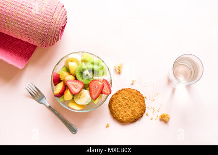 Sport und eine gesunde Lebensweise. Rosa Yoga Matte und Obst und Beeren Salat in der Schüssel und Vollkorn Cookie für Snack oder Frühstück auf Rosa pastell Bac Stockfoto
