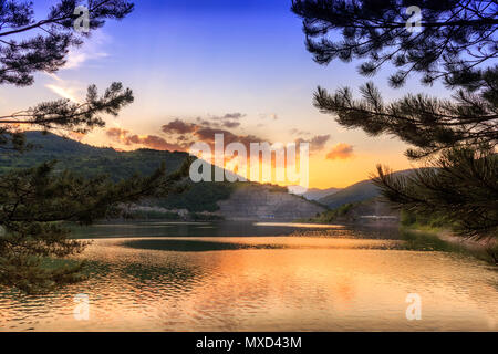 Pine Tree Branches framing reflektierenden See und goldenen Stunde dramatische Himmel mit dunklen, flauschige Wolken und tief oben Blau des Himmels Stockfoto