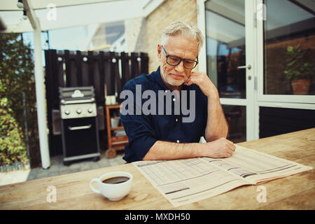 Lächelnd älterer Mann sitzen an seinem Tisch trinken eine Tasse Kaffee und der Lektüre der Seiten in einer Zeitung Stockfoto