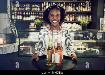 Lächelnden jungen afrikanischen Kellnerin in einem trendigen Restaurant Holding ein Tablett mit bunten Getränke Stockfoto