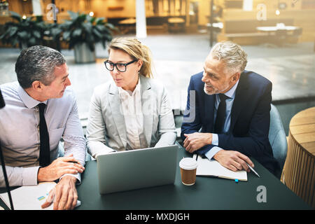 Drei ältere Führungskräfte lächelnd und miteinander sprechen, während Sie an einem Tisch in der Lobby eines Bürogebäudes sitzt Stockfoto