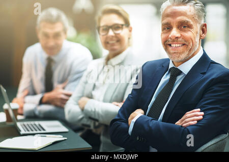 Zuversichtlich reife Geschäftsmann mit seinen Armen sitzen an einem Tisch in einem Büro gekreuzt mit lachenden Kollegen im Hintergrund Stockfoto