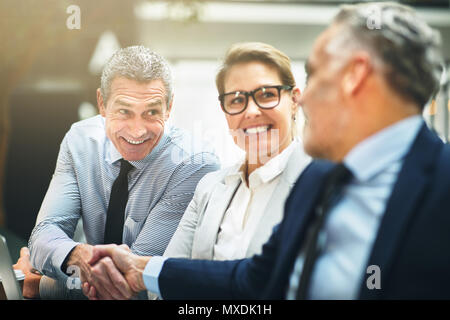 Zwei reife Geschäftsleute Lächeln und Händeschütteln zusammen mit einer weiblichen Kollegen dazu zu suchen, während an einem Tisch sitzen in einem Büro Stockfoto
