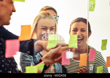 Lachend Gruppe von verschiedenen jungen Geschäftsleuten zusammen Brainstorming mit Haftnotizen auf eine Glaswand, während in einem modernen Büro stehend Stockfoto