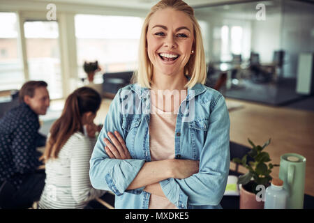 Lachende junge geschäftsfrau stehend, die Arme in einem Büro mit Kollegen an einem Tisch im Hintergrund arbeiten überschritten Stockfoto