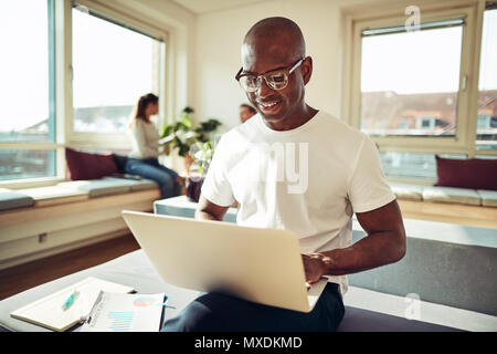 Lächelnd afrikanischen Geschäftsmann saß auf seinem Schreibtisch, die an einem Notebook arbeitet mit Kollegen zusammen gesprochen im Hintergrund Stockfoto