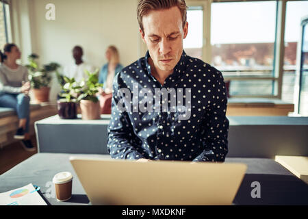 Konzentrierte sich junge Unternehmer online arbeiten mit einem Laptop sitzend auf seinem Schreibtisch in einem Büro mit Kollegen zusammen gesprochen im Hintergrund Stockfoto