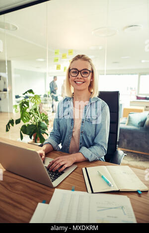 Leger gekleidete junge geschäftsfrau Brillen und selbstbewusst lächelnd, während Sie einen Laptop an Ihrem Schreibtisch in einem modernen Büro Stockfoto