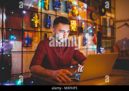 Eine junge Stattlichen kaukasischen Mann mit Bart und toothy Lächeln in einem rot kariertem Hemd ist die Arbeit hinter einem grauen Laptop an einem Holztisch sitzen. Die Hände auf der Tastatur. Abends an der Coffee Shop Stockfoto