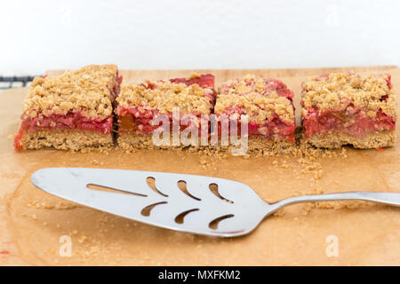 Scheiben von Rhabarber & Erdbeere bröckeln Bars, auf Backpapier mit einem silbernen cake Server angezeigt. Diese fruchtigen Leckereien sind ein gesunder Snack oder Dessert. Stockfoto