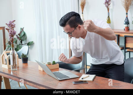 Asiatische legerer Mann stehen und bis Arm gute neue von mobilen am Laptop im Coffee Shop feiern, Erfolg Business Konzept, Arbeiten im Büro, zu Hause arbeiten Stockfoto