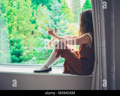 Eine junge Frau sitzt am Fenster an den Bäumen außerhalb während mit Ihrem Smart Phone auf der Suche Stockfoto