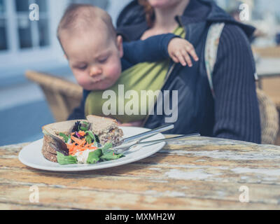 Eine Mutter mit einem Baby im Tragetuch sitzt an einem Tisch draußen mit einem Sandwich Stockfoto