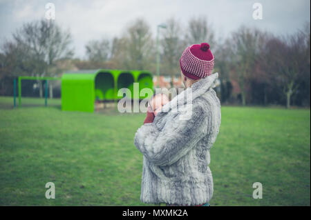 Eine junge Mutter steht auf dem Gras in einem Park im Winter mit ihrem Baby in einem Träger Schlinge Stockfoto