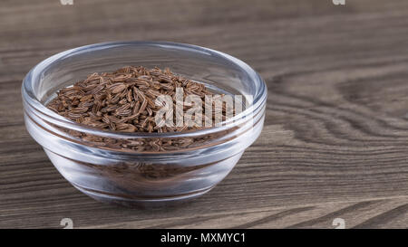 Kümmel in eine Glasschüssel auf Holz Hintergrund. Carum carvi. Schöne Nahaufnahmen der Haufen von Braun aromatischer Kümmel würzen. Charakteristischen Geschmack und Duft. Stockfoto