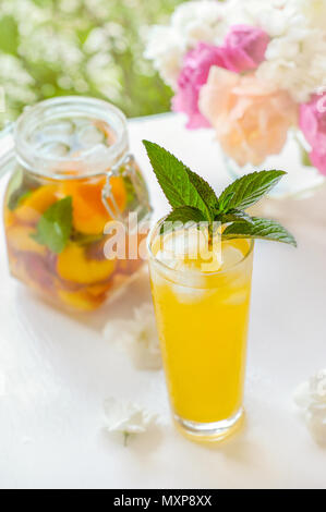 Krug mit Deckel mit Wasser, Detox mit Stücken von Pfirsich und Aprikose, mit Eis. Ein erfrischendes Getränk auf grünem Hintergrund mit einer gelben Rose. Stockfoto