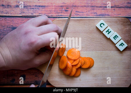 Koch ist Schneiden Möhre auf einer hölzernen Schneidebrett mit scharfen Messer und Wort Küchenchef mit Box Buchstaben geschrieben. Stockfoto