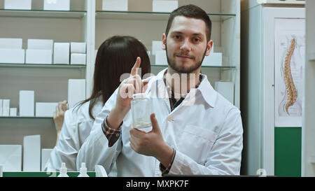 Junge malepharmacist Holding eine weiße Leere Flasche von Pillen, die Förderung der Medizin, während seine Kollegen arbeiten Stockfoto