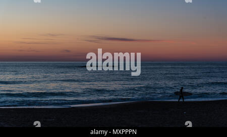 Ein Surfer zu Fuß entlang der Strand bei Sonnenuntergang Stockfoto