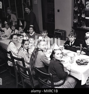 Historisches Bild der Kindergeburtstag, junge Kinder tragen Parteihüte sitzen zusammen an einem langen Tisch im Innenbereich ein Essen feiert Geburtstag des Kindes, Januar 1965, England, UK. Stockfoto