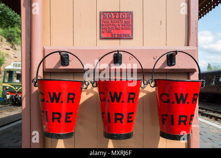 Nahaufnahme von drei roten GWR Notfeuerlöschpumpe Eimer auf Plattform im Vintage Steam Railway Station. Drei rote Eimer in einer Reihe. Trio der Eimer in der Sonne. Stockfoto