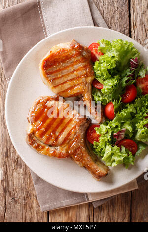 Gegrilltes Kotelett Schwein Honig lasiert, mit einem Salat von frischem Gemüse in der Nähe serviert auf einem Teller auf dem Tisch. Vertikal oben Ansicht von oben Stockfoto