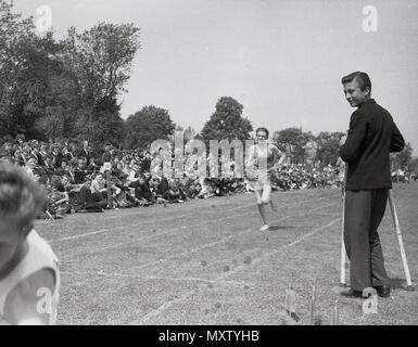 1960, historisches Bild der secondary school Jungen teil, die in einem laufenden Rennen auf einer inter-Schule county Sports Day, Dorset, England, UK. Der Sieger dieses Gras track Rennen scheint sich zu fallen nach der Ziellinie. Stockfoto