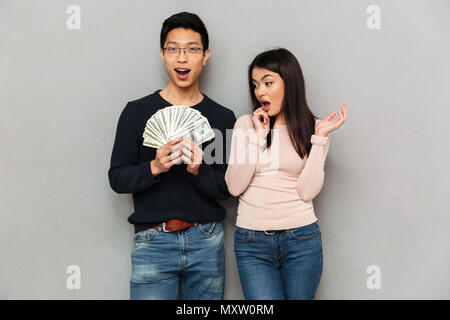 Bild der aufgeregten Jungen asiatischen Liebespaar stehend über graue Wand Hintergrund Geldhaltung isoliert. Stockfoto