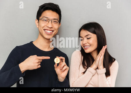 Bild von fröhlichen jungen asiatischen Liebespaar stehend über graue Wand Hintergrund holding Bitcoin isoliert. Stockfoto