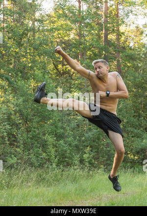 Mitte nach 40 s kaukasischen Mann barechested springen, Schlagen und Treten praktizieren Kampfkunst draußen im Wald Stockfoto