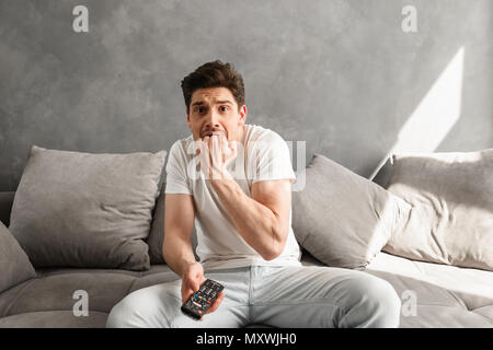 Foto von Angst, Mann beißt seine Faust in Angst beim Sitzen auf der Couch in grau Wohnung und halten Sie die Fernbedienung für den Fernseher in der Hand Stockfoto