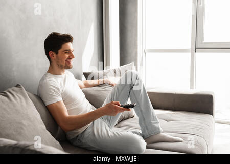 Bild im Profil der attraktiven brünetten Mann auf dem Sofa zu Hause sitzen und Fernsehen mit Fernbedienung in der Hand Stockfoto