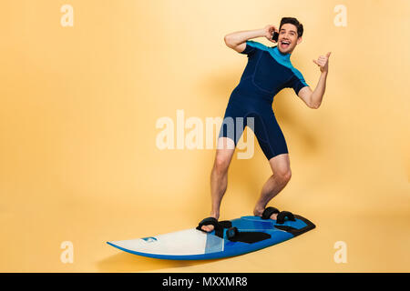 Foto einer aufgeregt, junger Mann im Badeanzug auf gelbem Hintergrund auf Surfen board Gespräch per Telefon isoliert gekleidet. Stockfoto