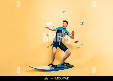 In voller Länge Porträt der glückliche junge Mann in Badeanzug Surfen auf einem Brett unter Geld gekleidet Dusche über der gelben Hintergrund isoliert