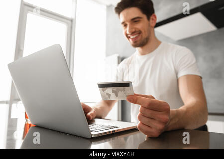 Foto von erfolgreichen Mann 30 s in Freizeitkleidung holding Kreditkarte und Zahlung auf Laptop in das moderne Apartment Stockfoto