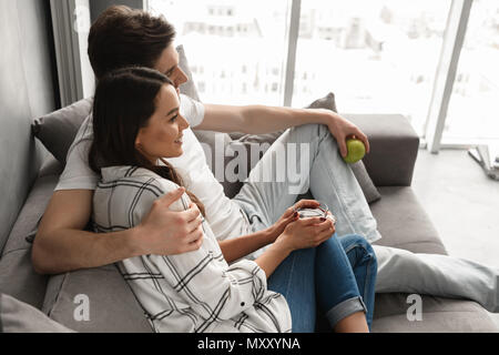 Foto von Paar schöne Mann und Frau sitzt auf einem Sofa in der Wohnung in der Nähe von Big Apple Fenster beim Essen und Tee trinken. Stockfoto