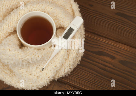 Eine Teetasse in einem weichen, flauschigen gestrickter Schal gewickelt, ein Thermometer auf einer hölzernen Hintergrund - das Konzept der saisonalen Herbst Krankheiten, Erkältungen, Grippe Behandlung Stockfoto