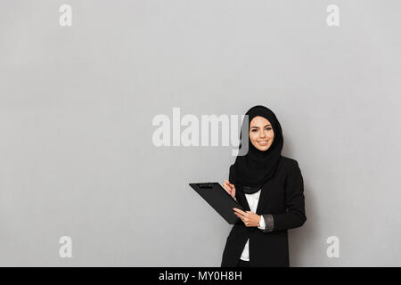 Portrait von muslimischen lächelnde Frau 20 s in Schwarz traditionelle Kleidung auf Kamera halten Zwischenablage mit Dokumenten über Grau isoliert backgro Stockfoto