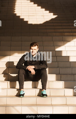Porträt eines lächelnden jungen Sportler der Ohrhörer ruht beim Sitzen auf der Treppe im Freien Stockfoto