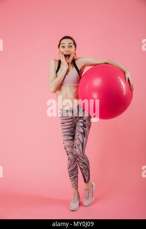 Volle Länge Bild von Schockiert glücklich Sportlerin posiert mit Fitness Ball, während ihre Wange berühren, und wenn man die Kamera über rosa Hintergrund Stockfoto