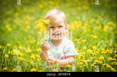 Schöne glückliche kleine Baby Mädchen sitzen auf eine grüne Wiese mit gelben Blumen Löwenzahn Stockfoto