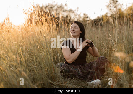 Das Porträt einer Frau im Gras sitzen und ihr Haar zu tun. Eine junge Frau genießen Haare flechten in der Natur bei Sonnenuntergang. Stockfoto