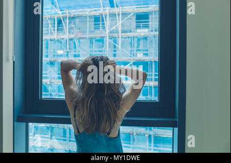 Eine junge Frau, die durch das Fenster in ein Hochhaus ist genervt von den Bauarbeiten außerhalb Stockfoto