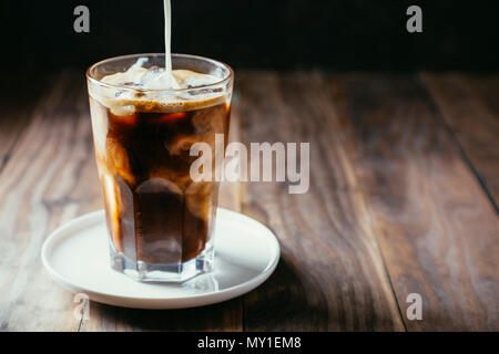 Gießen Milch in einen gefrorenen Kaffee in einem hohen Glas mit einem Stroh auf einem rustikalen Holztisch Stockfoto
