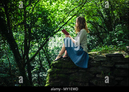 Eine junge Frau sitzt auf einer Wand in den Wald und ist ein Buch lesen Stockfoto