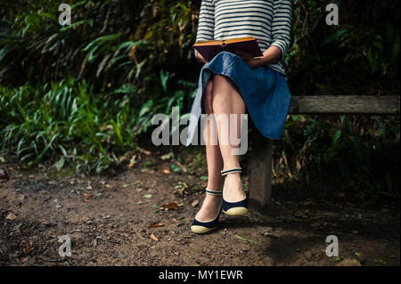 Eine junge Frau sitzt auf einer Bank im Wald und ist ein Buch lesen Stockfoto